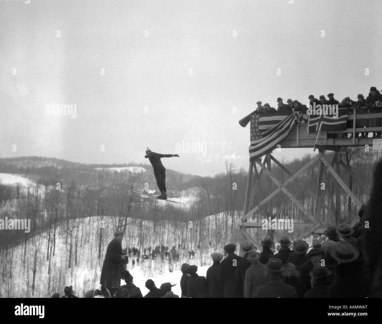1930s Uomo salto con gli sci a metà in aria la folla di spettatori GRAND STAND drappeggiati con bandiere e BUNTING INVERNO paesaggio innevato Foto Stock
