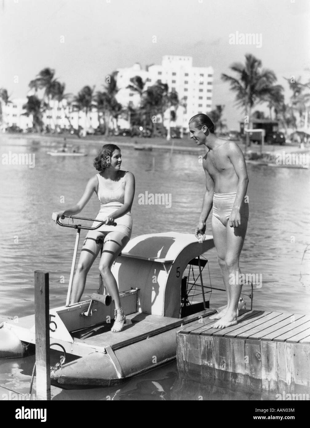 1930s GIOVANE UOMO DONNA COSTUME DA BAGNO Vestiti donna seduta sulla barca del pedale uomo in piedi sul dock Foto Stock