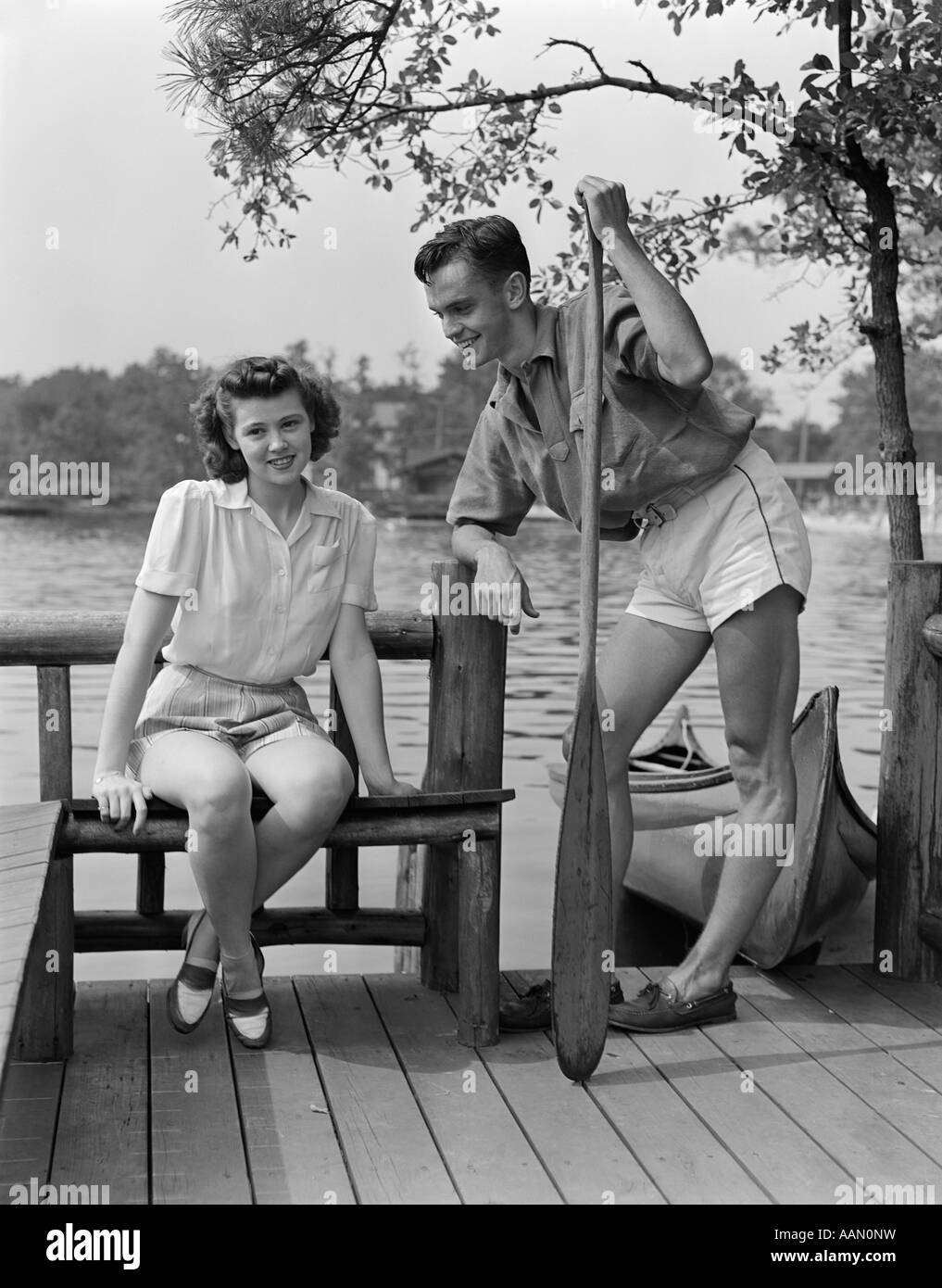 1940s l uomo e la donna sul dock dal lago di donna seduta MAN STANDING HOLDING OAR CANOA IN BACKGROUND FLIRT ESTIVO Foto Stock