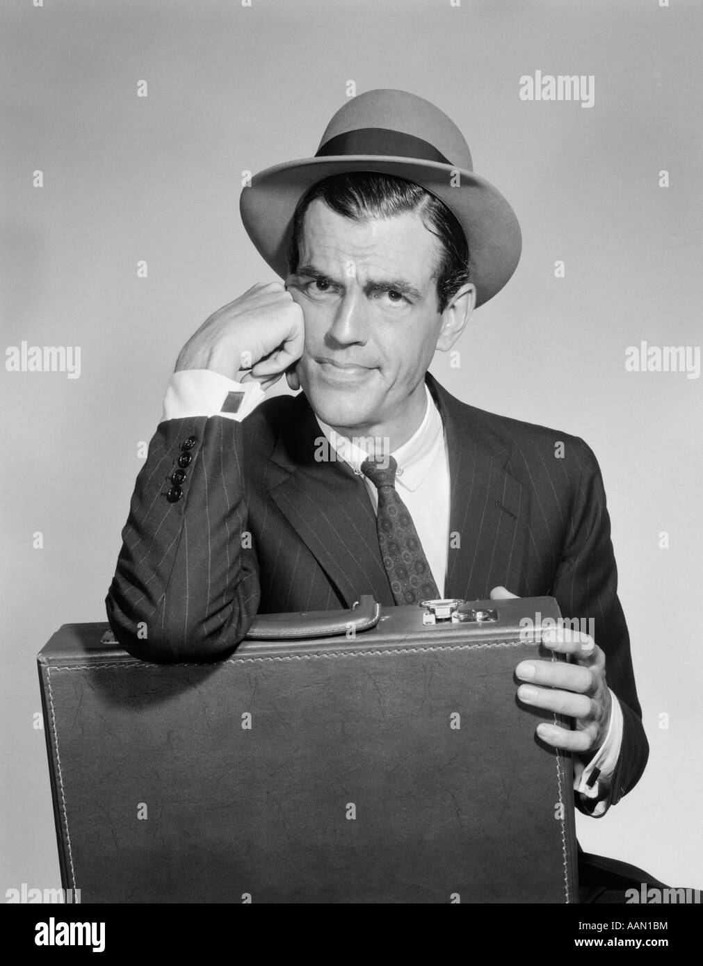 Anni Cinquanta venditore valigetta di contenimento in giro in appoggio il gomito sul caso faccia di appoggio sul lato preoccupato confuso arrabbiato indossando HAT Foto Stock