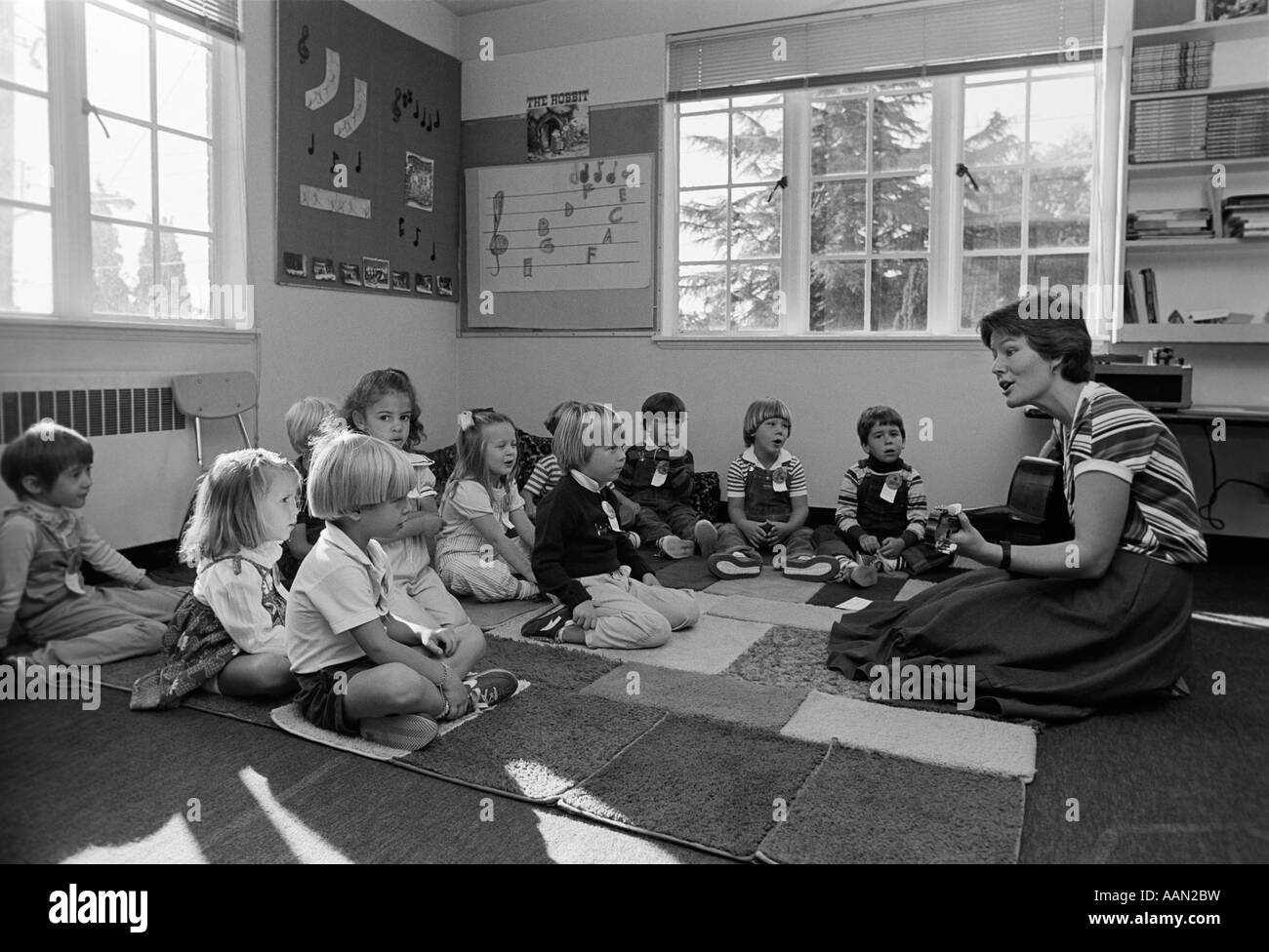Anni ottanta GRADE SCHOOL insegnante seduto sul pavimento con gli studenti a suonare la chitarra e cantare Foto Stock