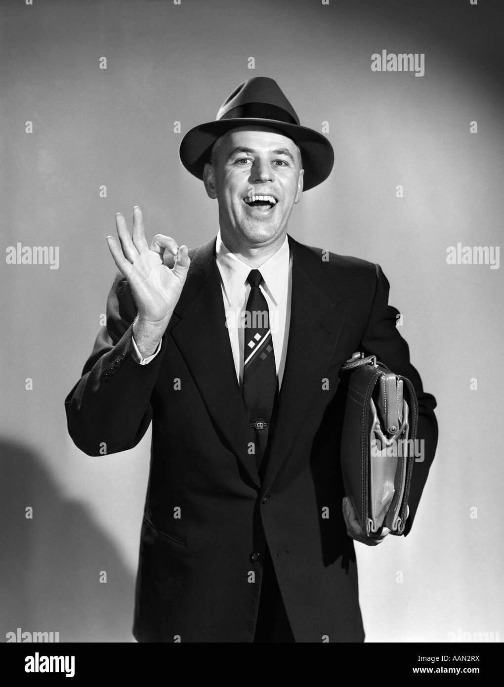 Anni Cinquanta venditore uomo rendendo OKAY segno con il pollice e il dito indice VALIGETTA TRASPORTO indossare tuta TIE HAT Foto Stock