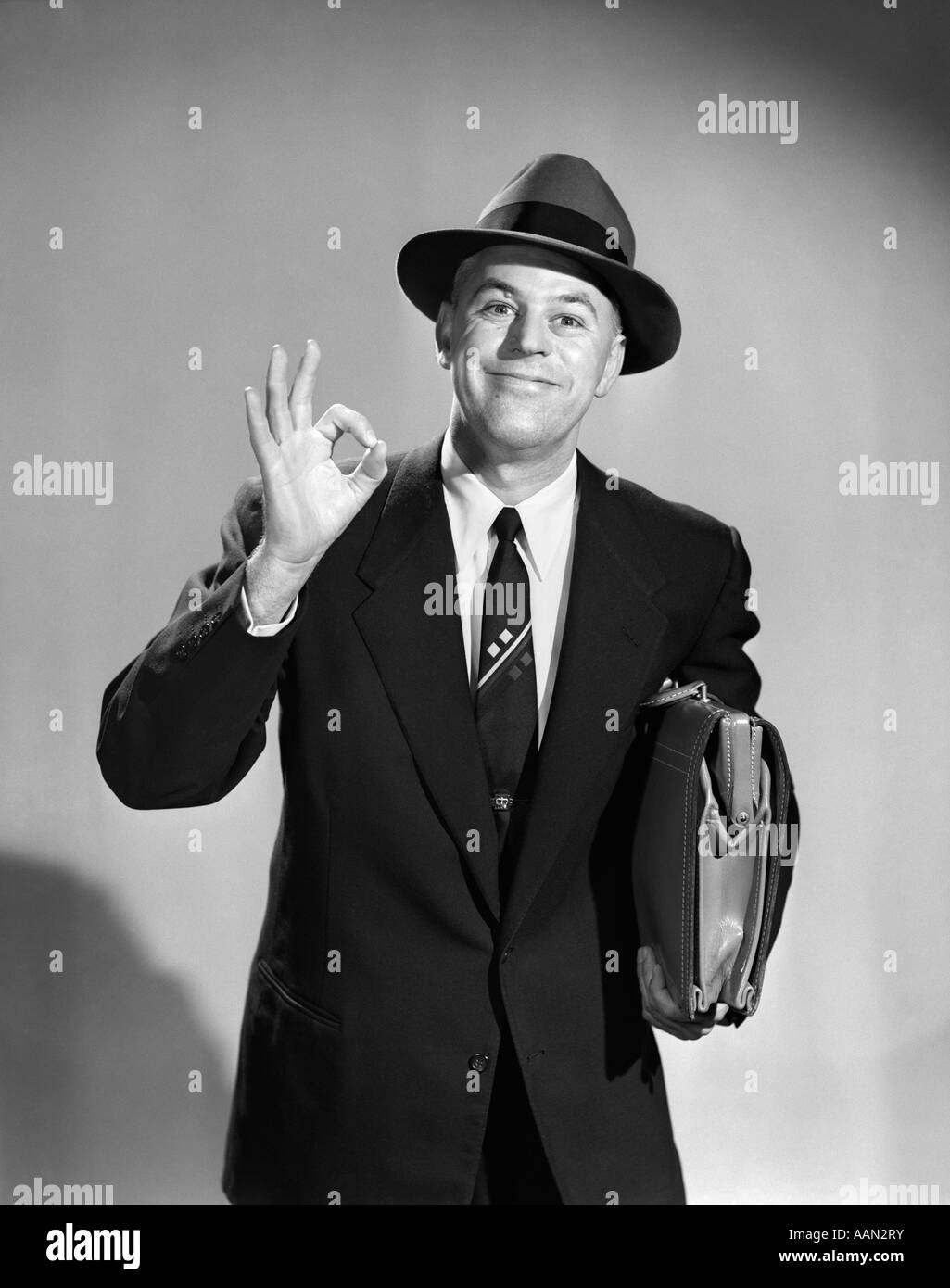 Anni Cinquanta uomo imprenditore venditore rendendo OKAY SEGNO CON UNA MANO valigetta porta indossando HAT guardando la fotocamera Foto Stock