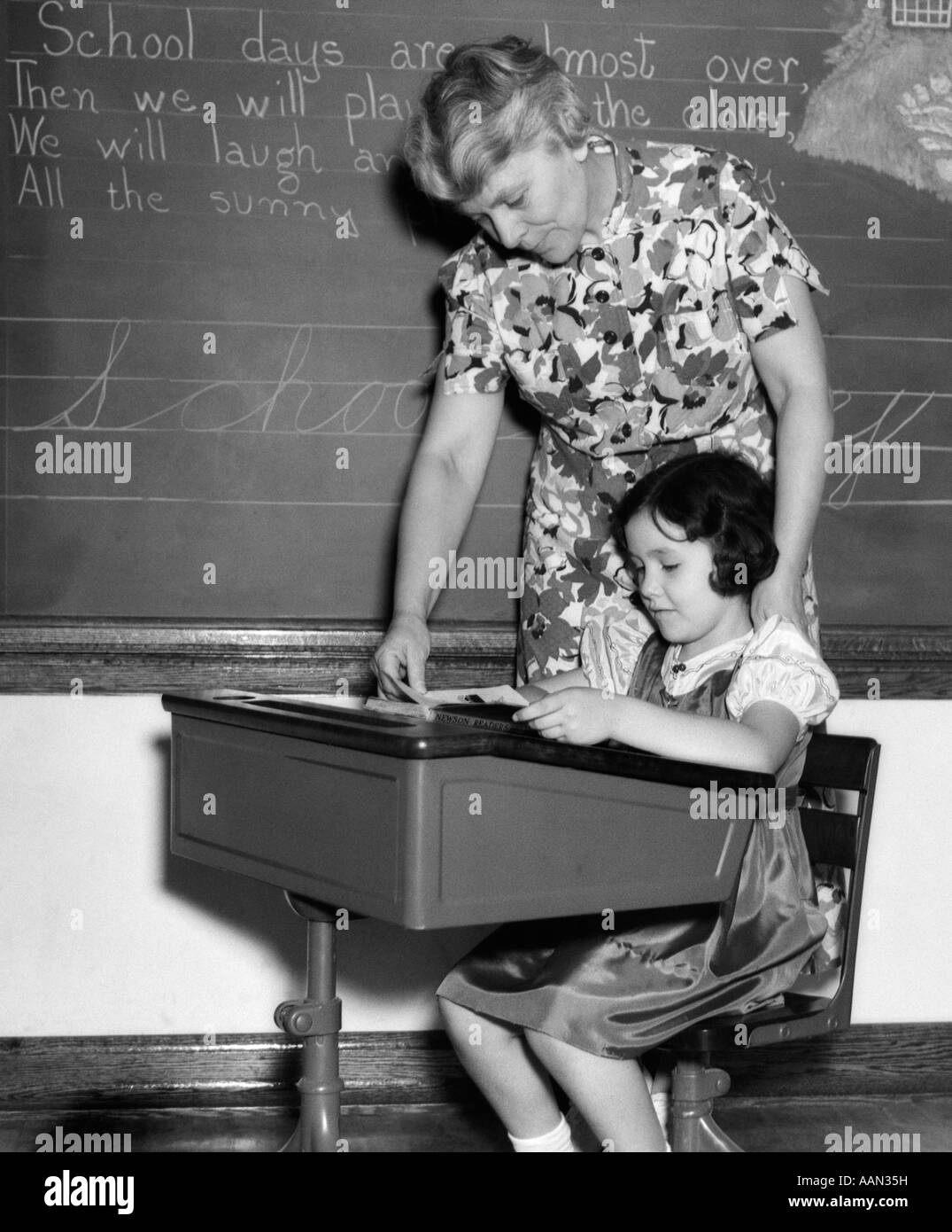 1930 Insegnante FEMMINILE NELLA PARTE ANTERIORE DI BLACKBOARD aiutando una studentessa al desk LIBRO DI LETTURA Foto Stock