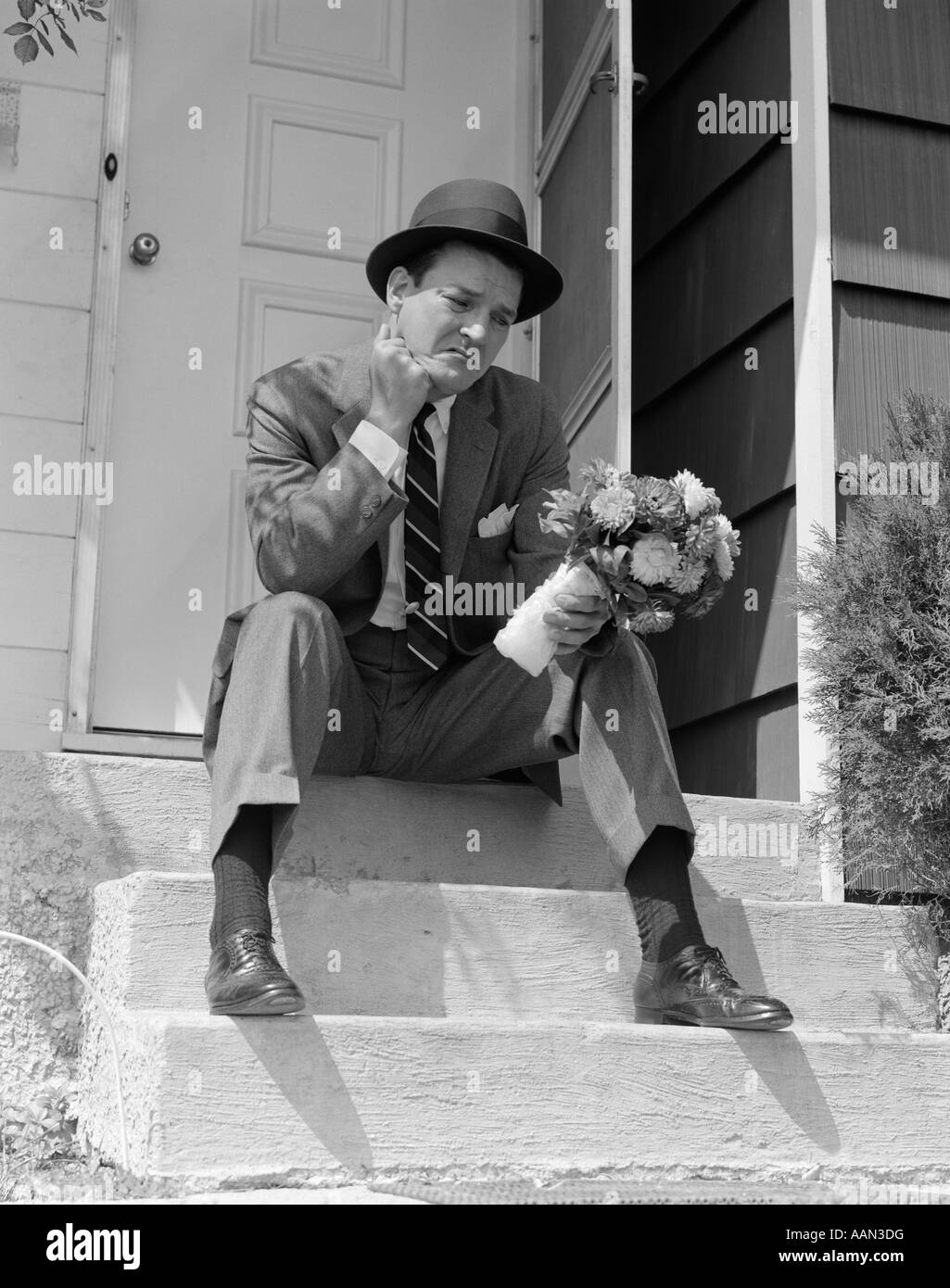 Anni Cinquanta UOMO IN TUTA E HAT HOLDING fiori seduto fuori porta anteriore sui passi cercando triste Foto Stock