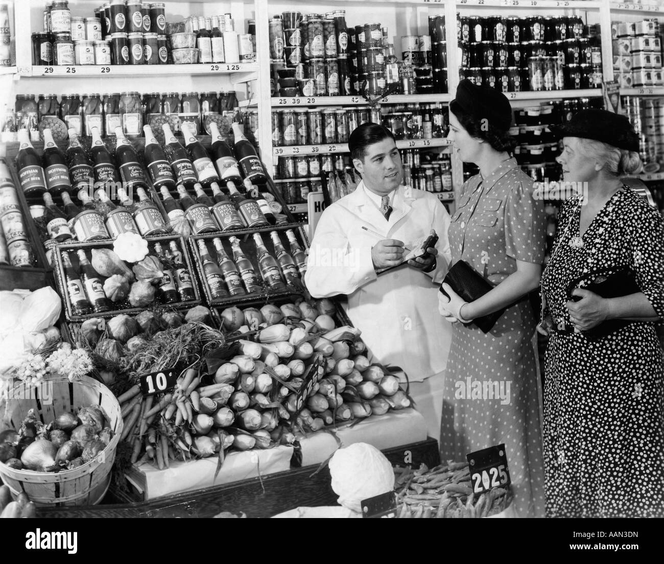 1930s grocer nel mantello bianco AZIENDA PAD e matita per prendere gli ordini verso il basso da due donne amanti dello shopping Foto Stock