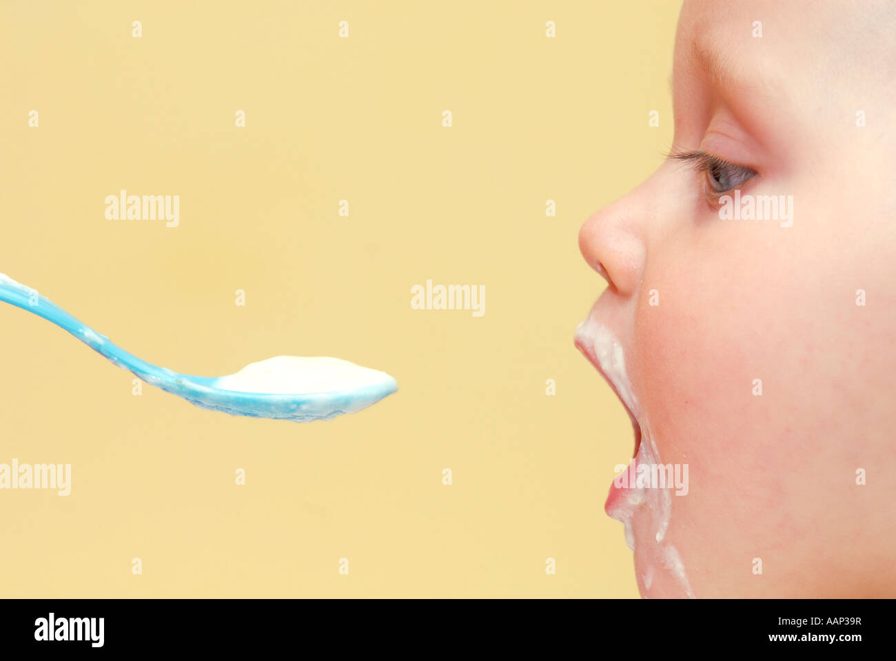 Bambini baby boy in sedia alta mangiare yogurt con cucchiaio blu, sfondo giallo, dettaglio close up, vista laterale, profilo Foto Stock