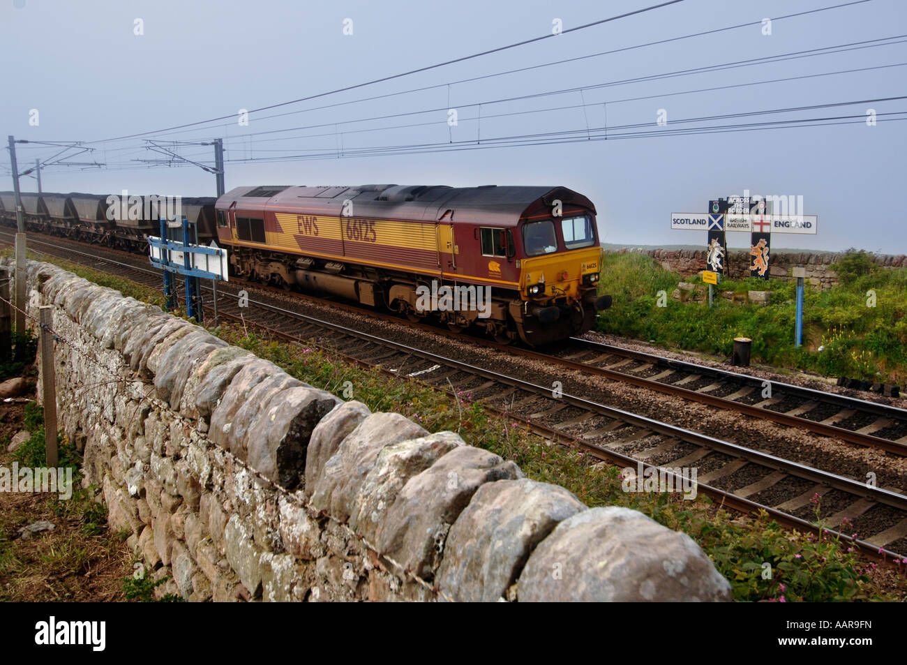 Un EWS freight attraversa il confine dalla Scozia in Inghilterra su un treno di carbone passando un vecchio British Railways segno con bandiere Foto Stock