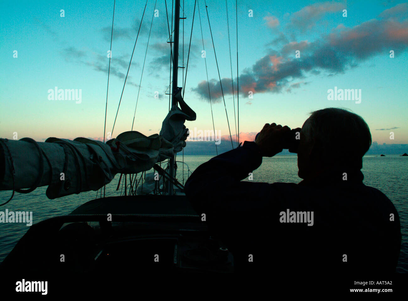 A sailor on an ocean passage searches the horizon with binoculars from the deck of his sailboat. Perhaps he looks for land Foto Stock