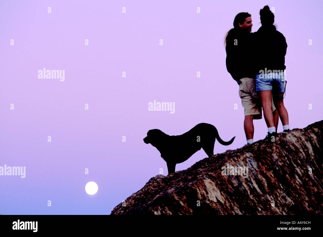 Amare giovane sulla cima di una montagna al tramonto a guardare la luna piena di salita con il loro cane fissando la luna Foto Stock