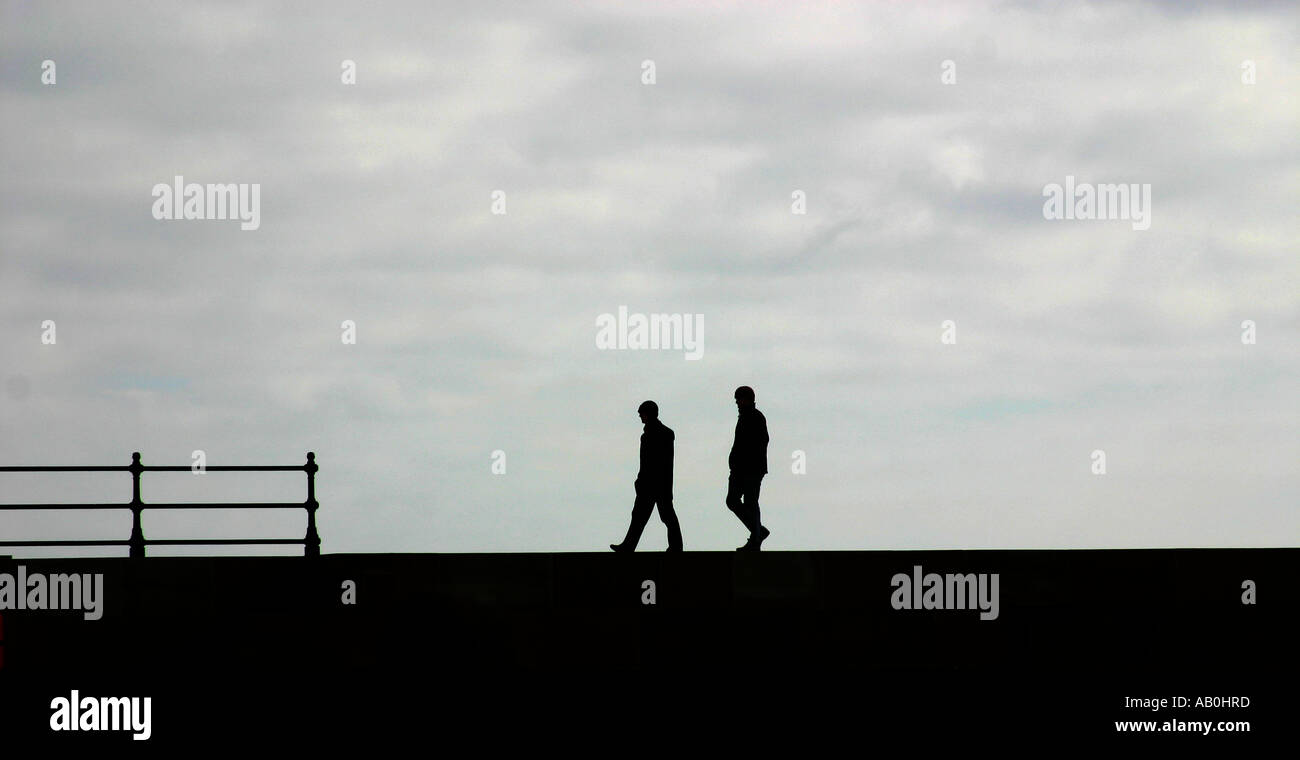 Due persone a piedi lungo la parte superiore di una parete del porto in silhouette. Foto Stock