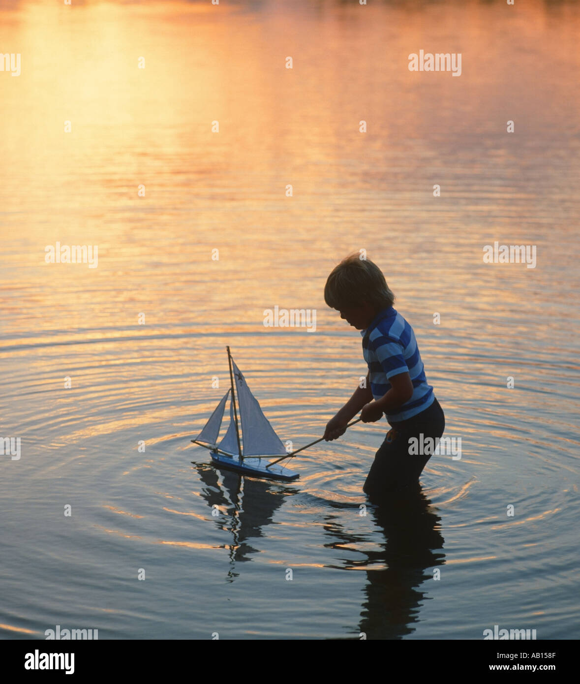 Giovane ragazzo giocando nel lago con il giocattolo in barca a vela al tramonto Foto Stock