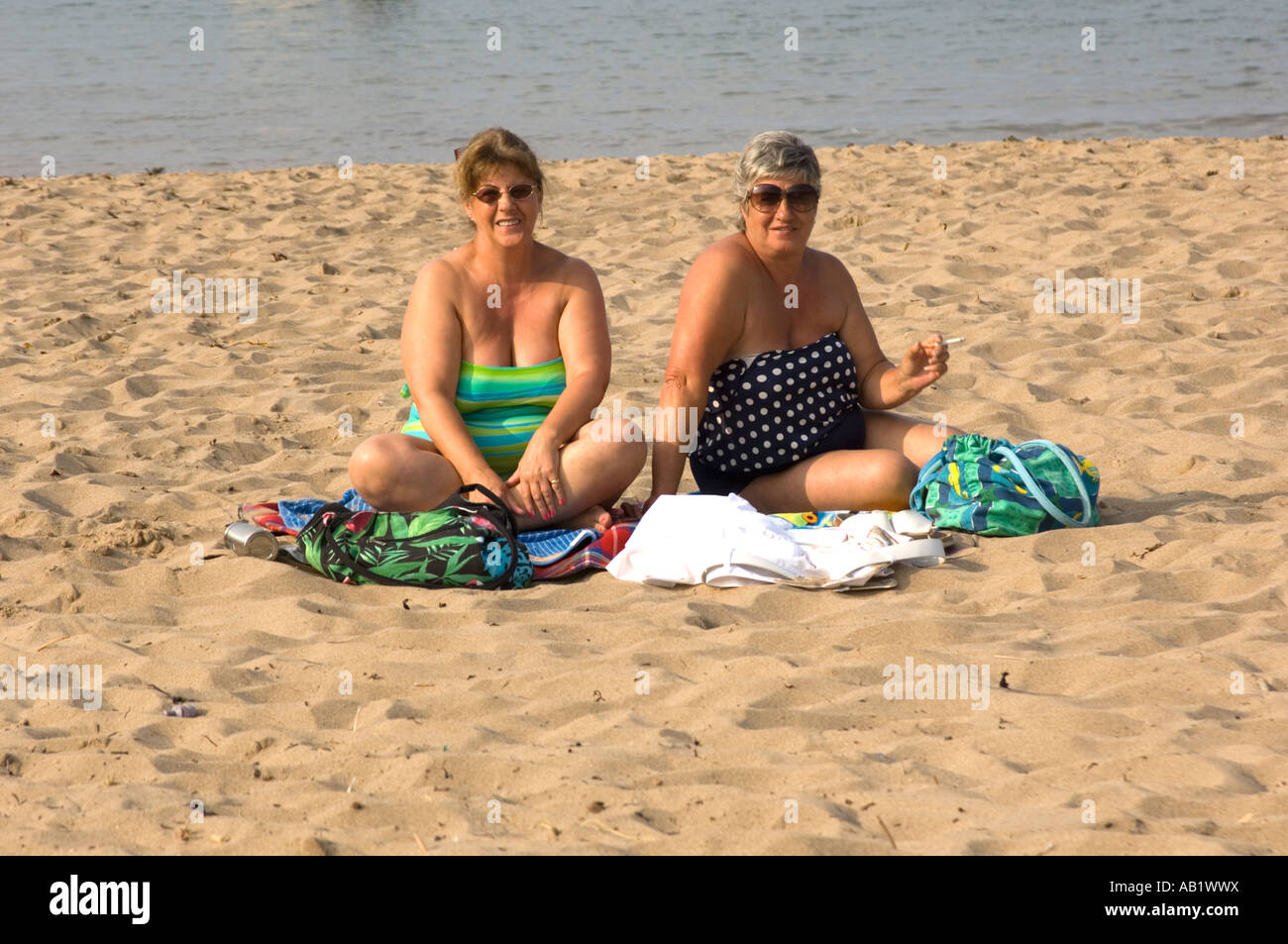 Due donne in sovrappeso (due fat ladies) su di una spiaggia di sabbia con una sigaretta nella mano sinistra guardando la telecamera, pomeriggio estivo Foto Stock