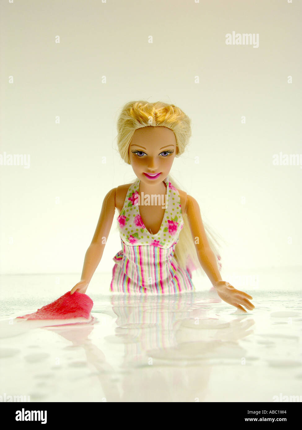 Una bionda bambola Barbie si inginocchia in un abito di fiori prima di sfondo bianco su un terreno bianco con ornamenti in acqua e un panno per pulire la Foto Stock