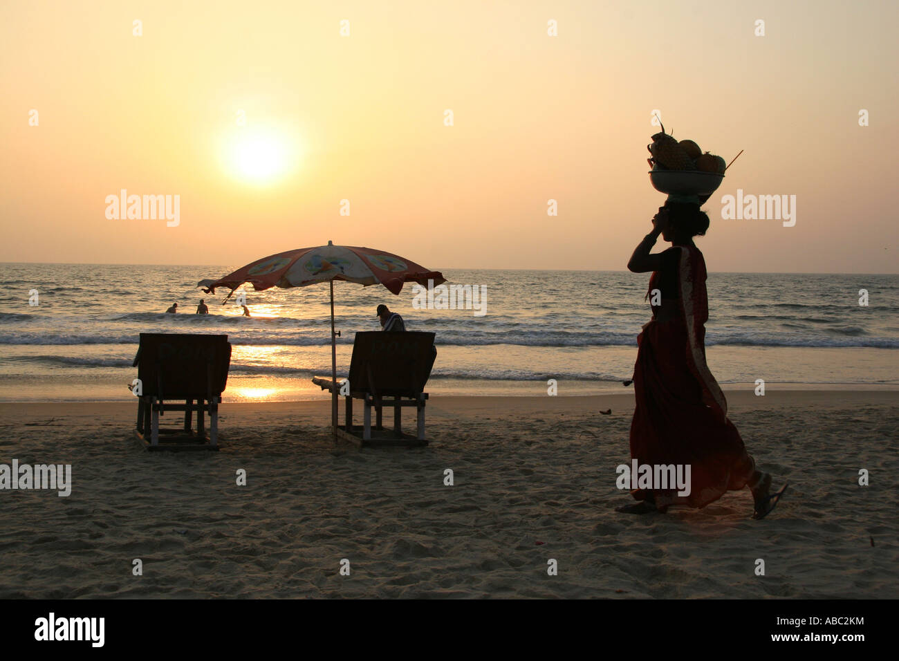 Tramonto su una spiaggia nella parte sud di Goa. Una donna Indiana che porta un cesto di frutta si stagliano contro il sole di setting. Foto Stock