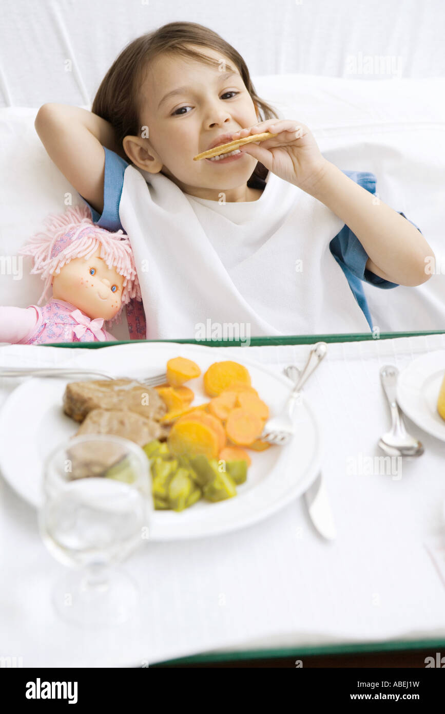 Ragazza distesa nel letto di ospedale con il vassoio per alimenti di fronte a lei, mangiare e cookie Foto Stock