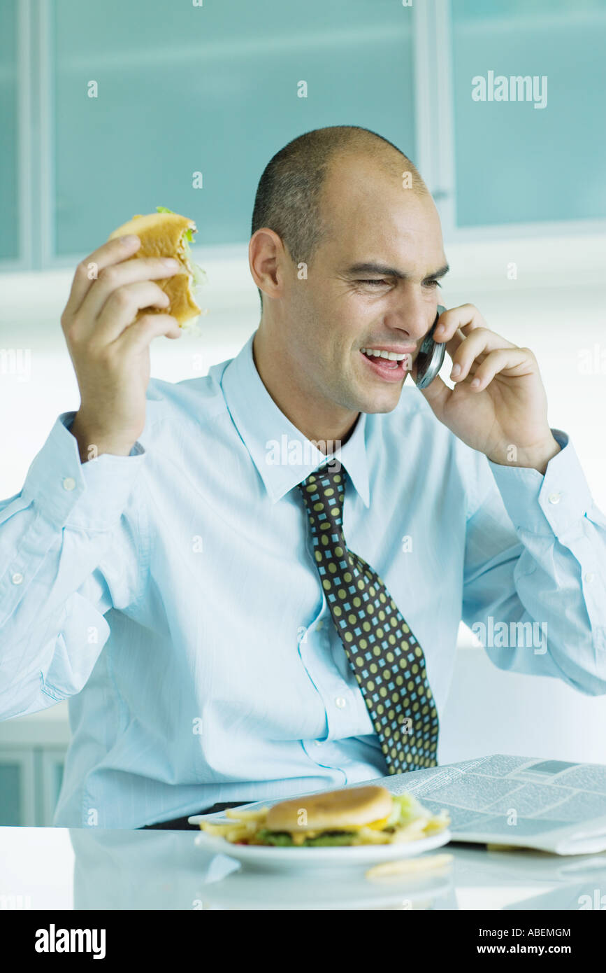 Uomo seduto a tavola a mangiare hamburger, leggendo il giornale e utilizzando il telefono cellulare Foto Stock