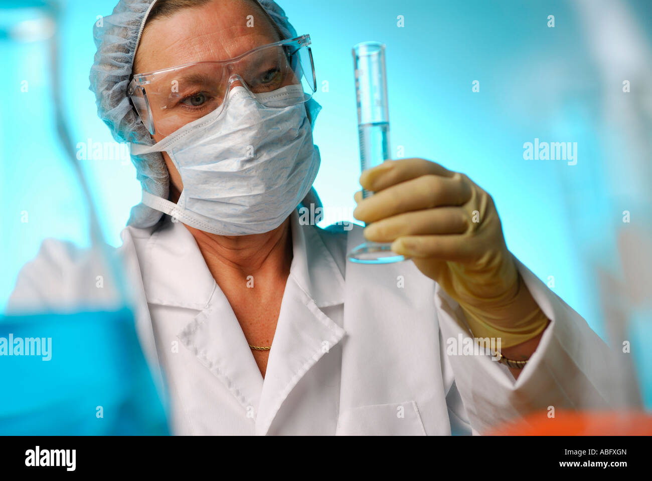 Femmina tecnico di laboratorio con occhiali di sicurezza guanti maschera e retina per capelli il controllo volume del liquido nel cilindro graduato su sfondo blu Foto Stock