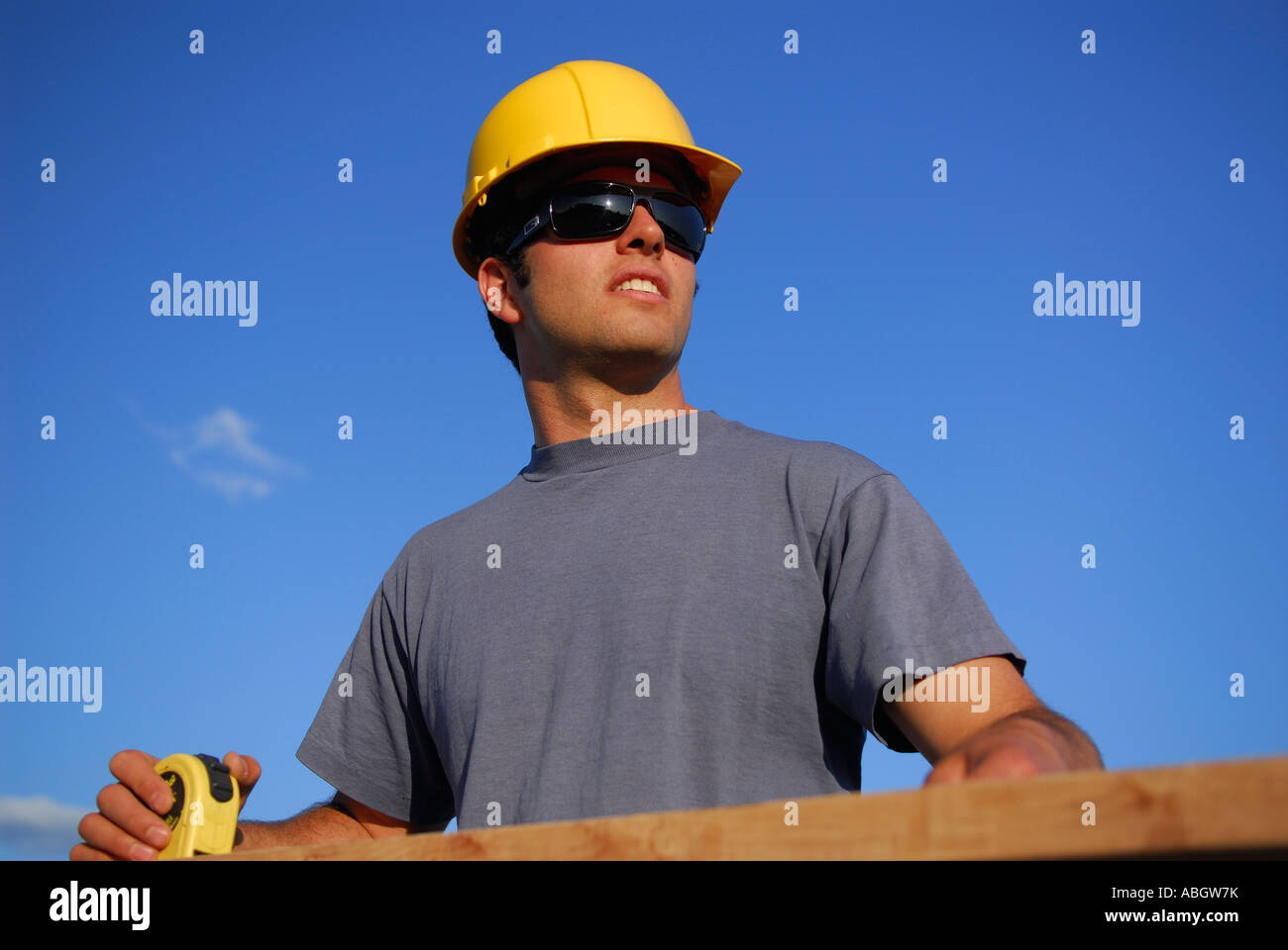 Operaio edile falegname in hard hat cercando durante la misurazione asse di legno con nastro di misurazione contro il cielo blu Foto Stock