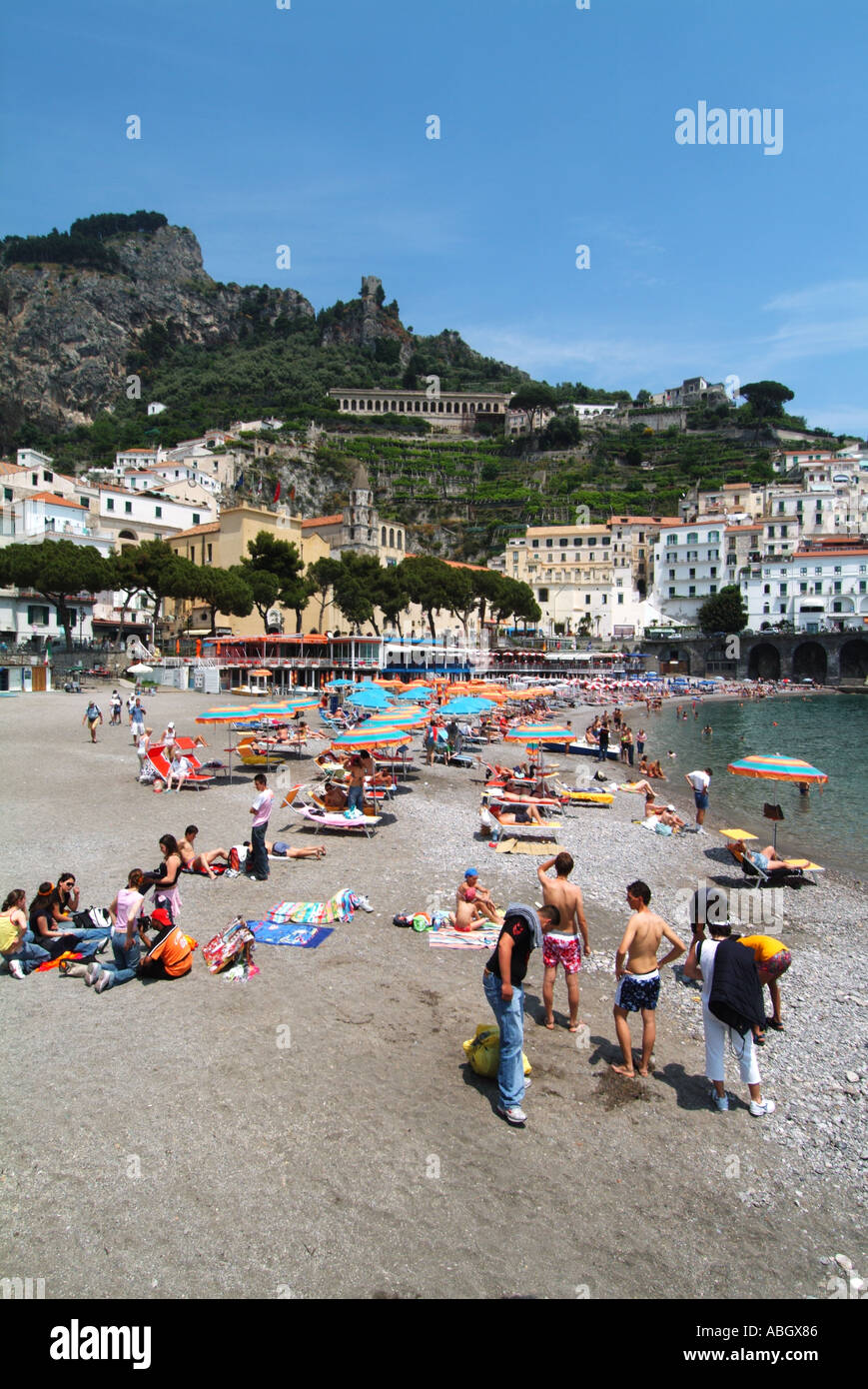 Turisti e gente del posto estate prendere il sole in pietra alla fine della spiaggia di Amalfi, vista sulla spiaggia costiera lungo il bordo delle acque fino agli ombrelloni Campania Salerno Italia e UE Foto Stock