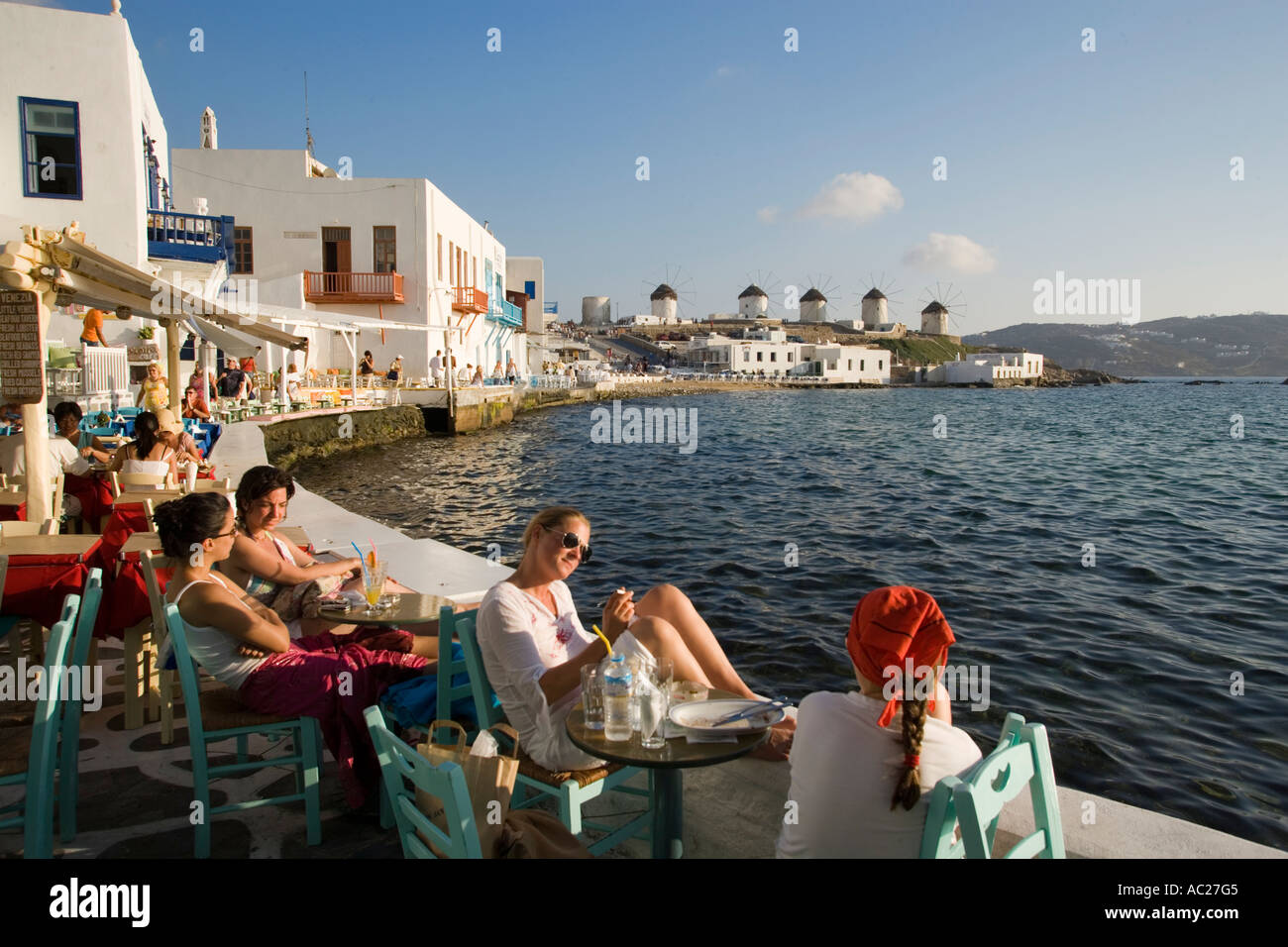 La gente seduta in ristoranti e bar direttamente in mare di mulini a vento in background la piccola Venezia città di Mykonos Mykonos Grecia Foto Stock