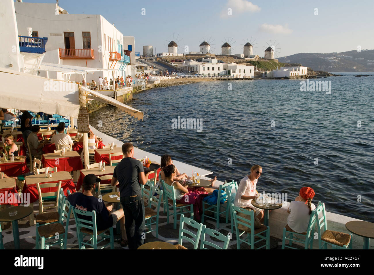 La gente seduta in ristoranti e bar direttamente in mare di mulini a vento in background la piccola Venezia città di Mykonos Mykonos Grecia Foto Stock