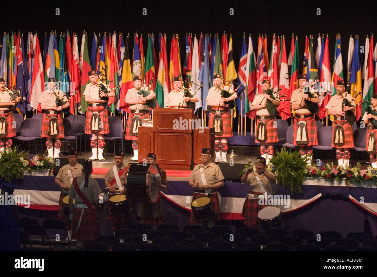 Gli uomini in uniformi scozzese riproduzione di cornamuse e percussioni in una banda di cornamuse esibirsi sul palco di fronte a bandiere internazionali Foto Stock