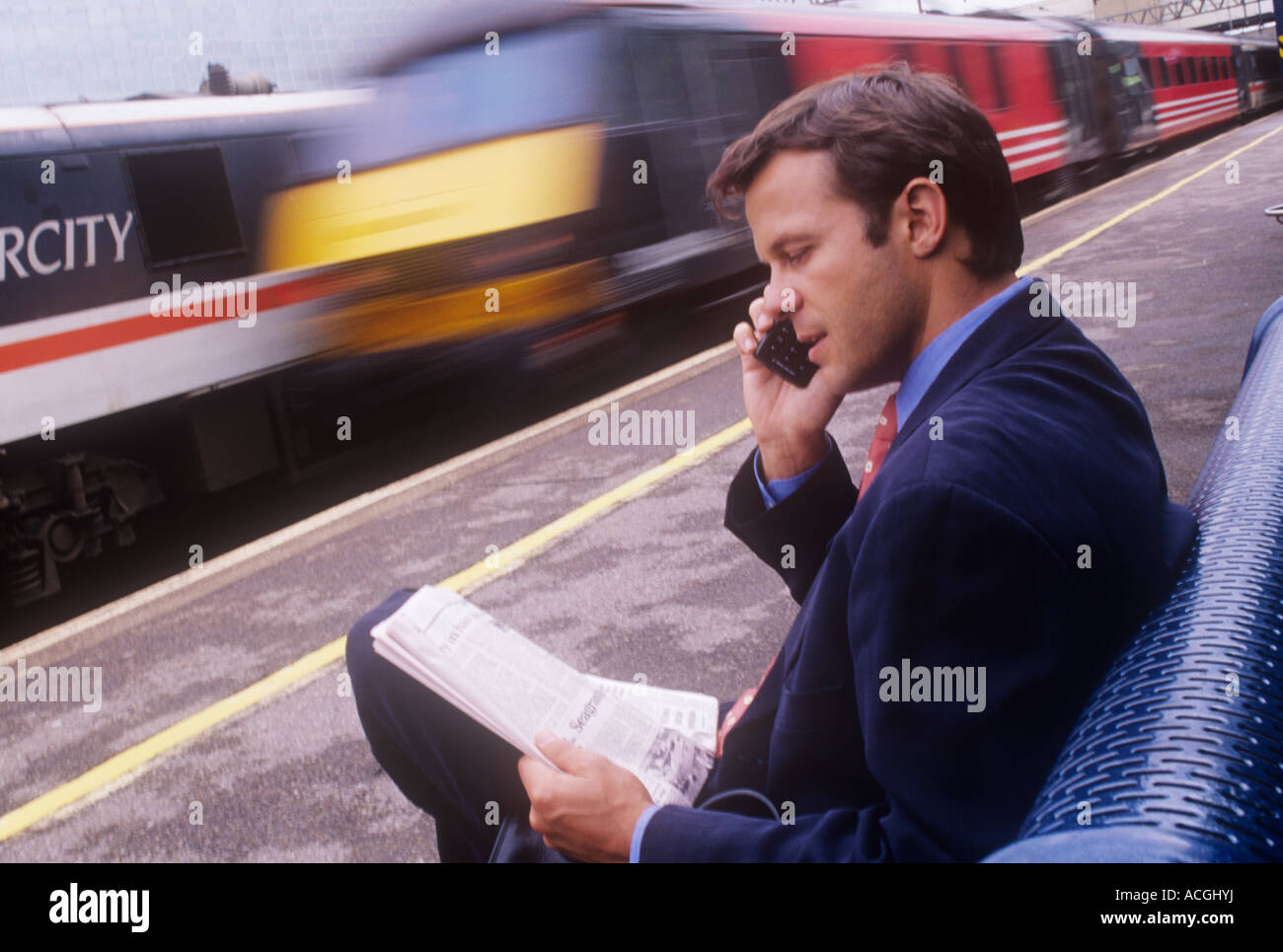 Uomo d'affari seduto sul banco della piattaforma della stazione ferroviaria, leggendo un giornale e parlando sul suo telefono cellulare, con Virgin treno che passa con la velocità b Foto Stock