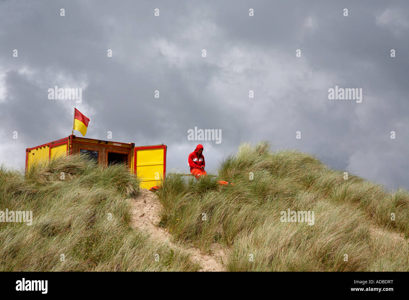 La vita di guardia di abbigliamento rossa si trova sulla parte superiore della zona erbosa guardando curracloe spiaggia come temporale si sposta in Foto Stock