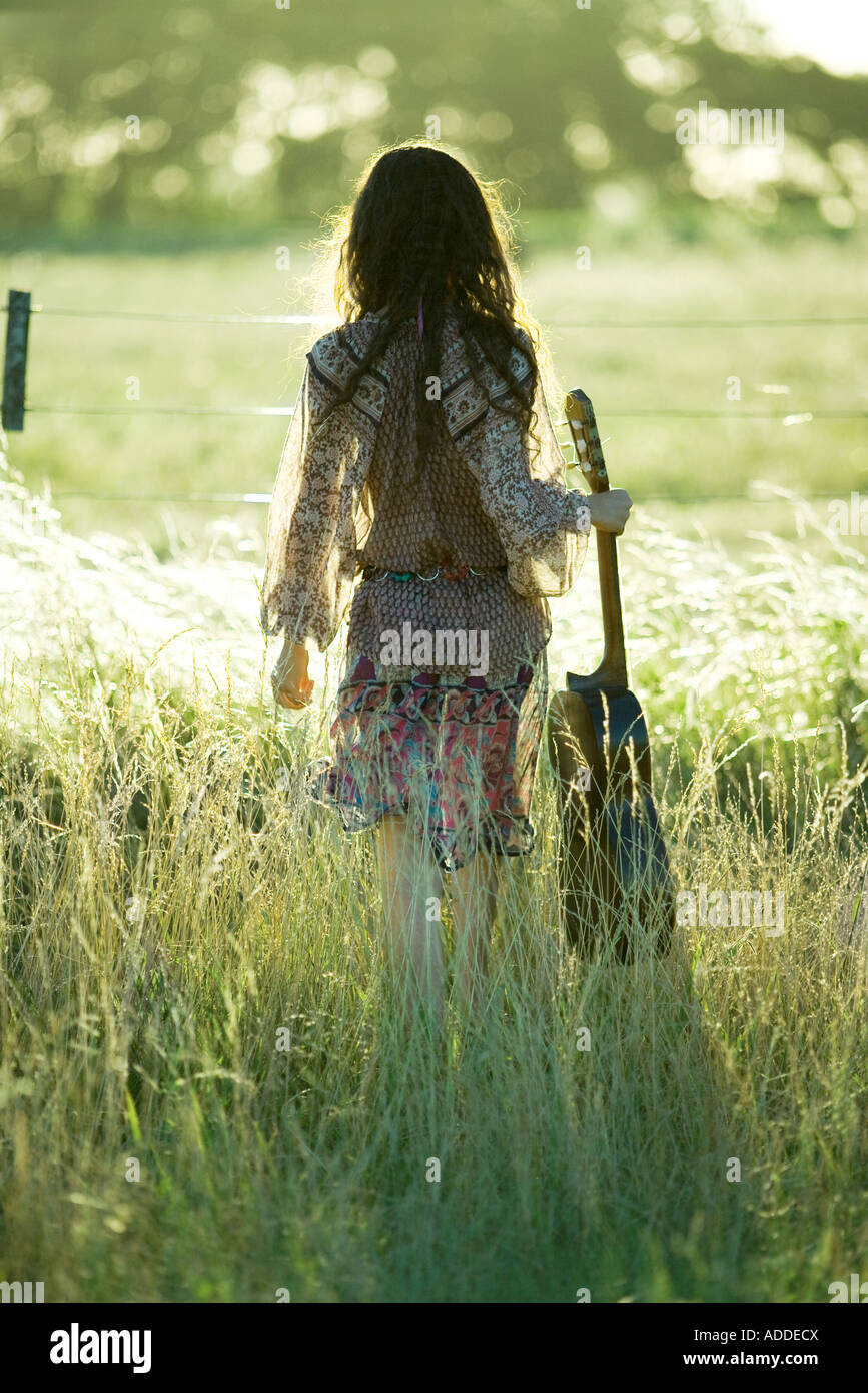 Giovane donna hippie tenendo la chitarra in piedi in campo, vista posteriore Foto Stock