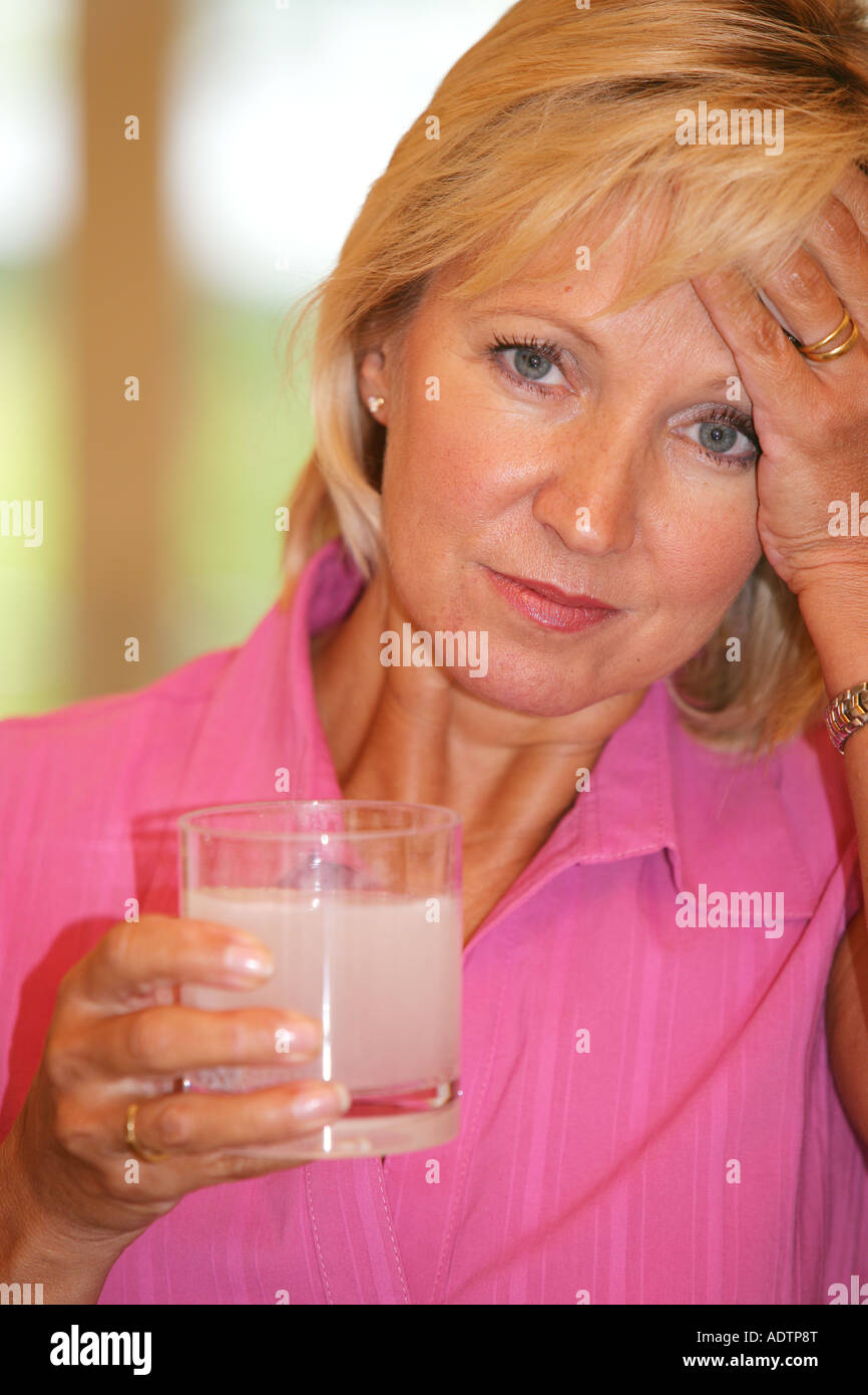 Donna matura senso di malessere con un mal di testa tenendo solubile antidolorifiche compresse in un bicchiere tenendo la testa indossando un Top rosa Foto Stock