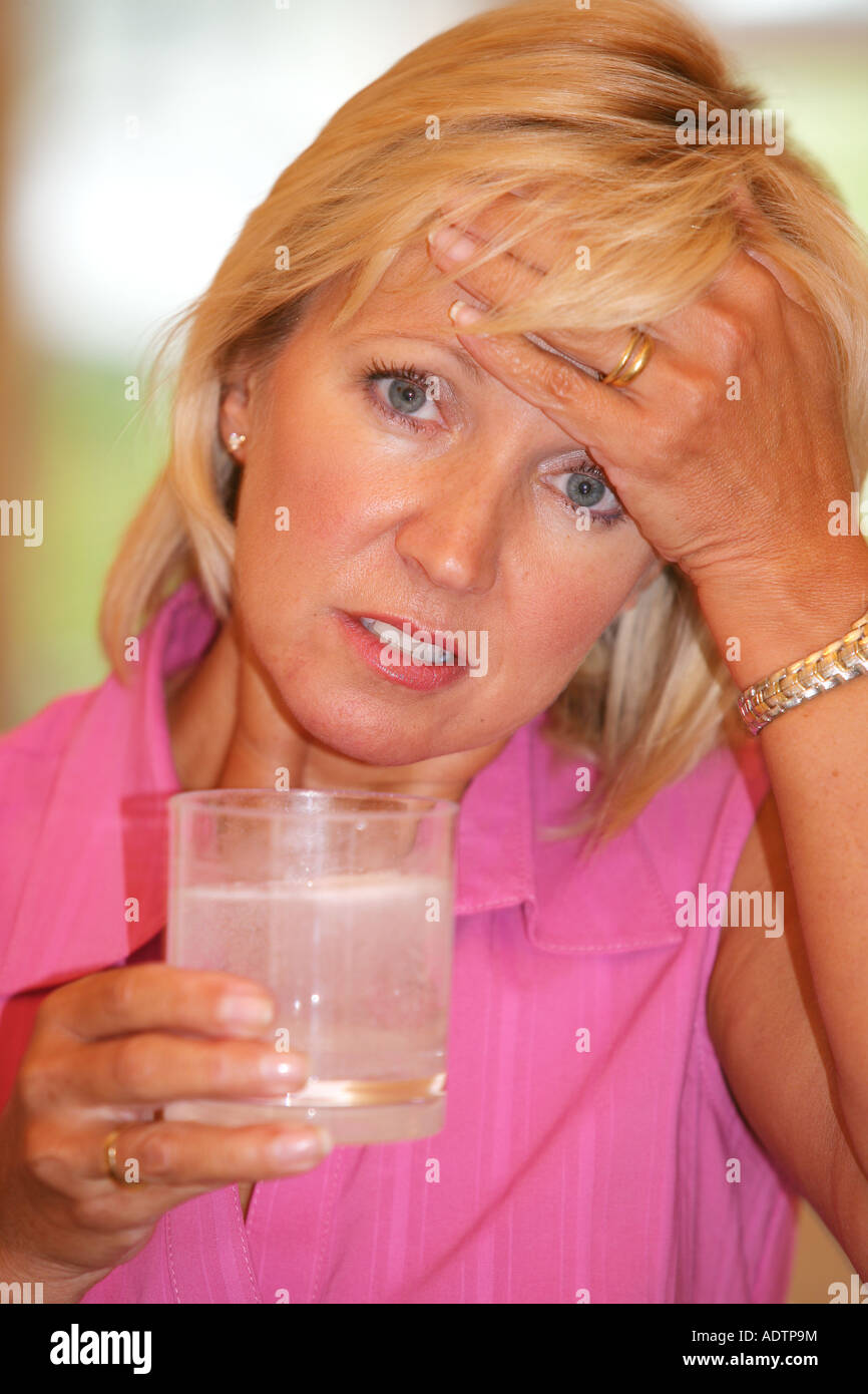 Donna matura senso di malessere con un mal di testa tenendo solubile antidolorifiche compresse in un bicchiere tenendo la testa indossando un Top rosa Foto Stock