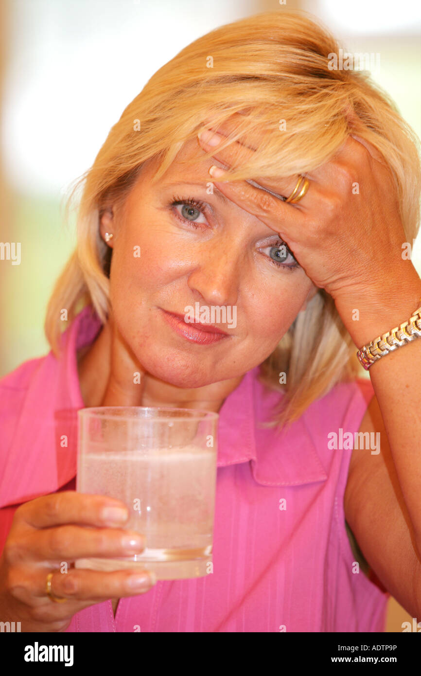 Donna matura senso di malessere con un mal di testa tenendo solubile antidolorifiche compresse in un bicchiere tenendo la testa indossando un Top rosa Foto Stock