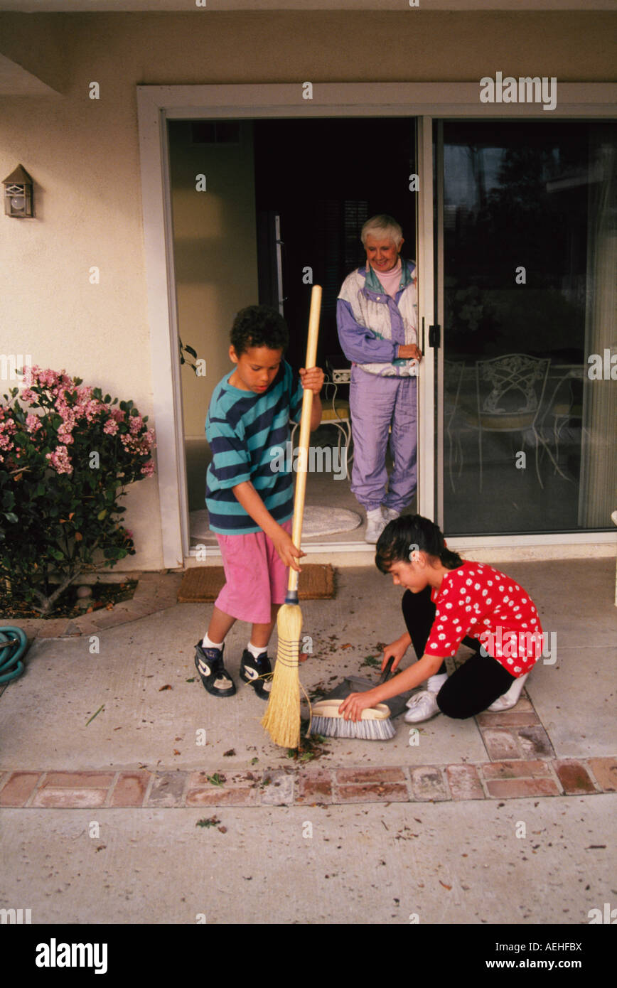 Diversi bambino aiutando un altro ragazzo ragazza 6-8 anno vecchio donna anziana riordino spazzare spazza sweep patio porticato servire volontario aiuta gli anziani Foto Stock
