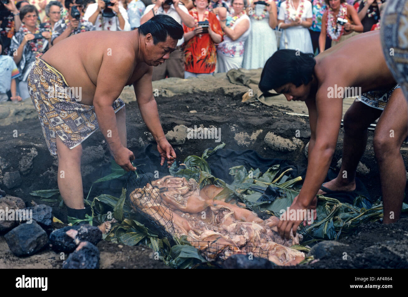 I visitatori osservano due grandi uomini locali scoprire maiale arrostito da pietre calde nella fossa di massa a un luau tradizionale festa in Kona Hawaii Foto Stock