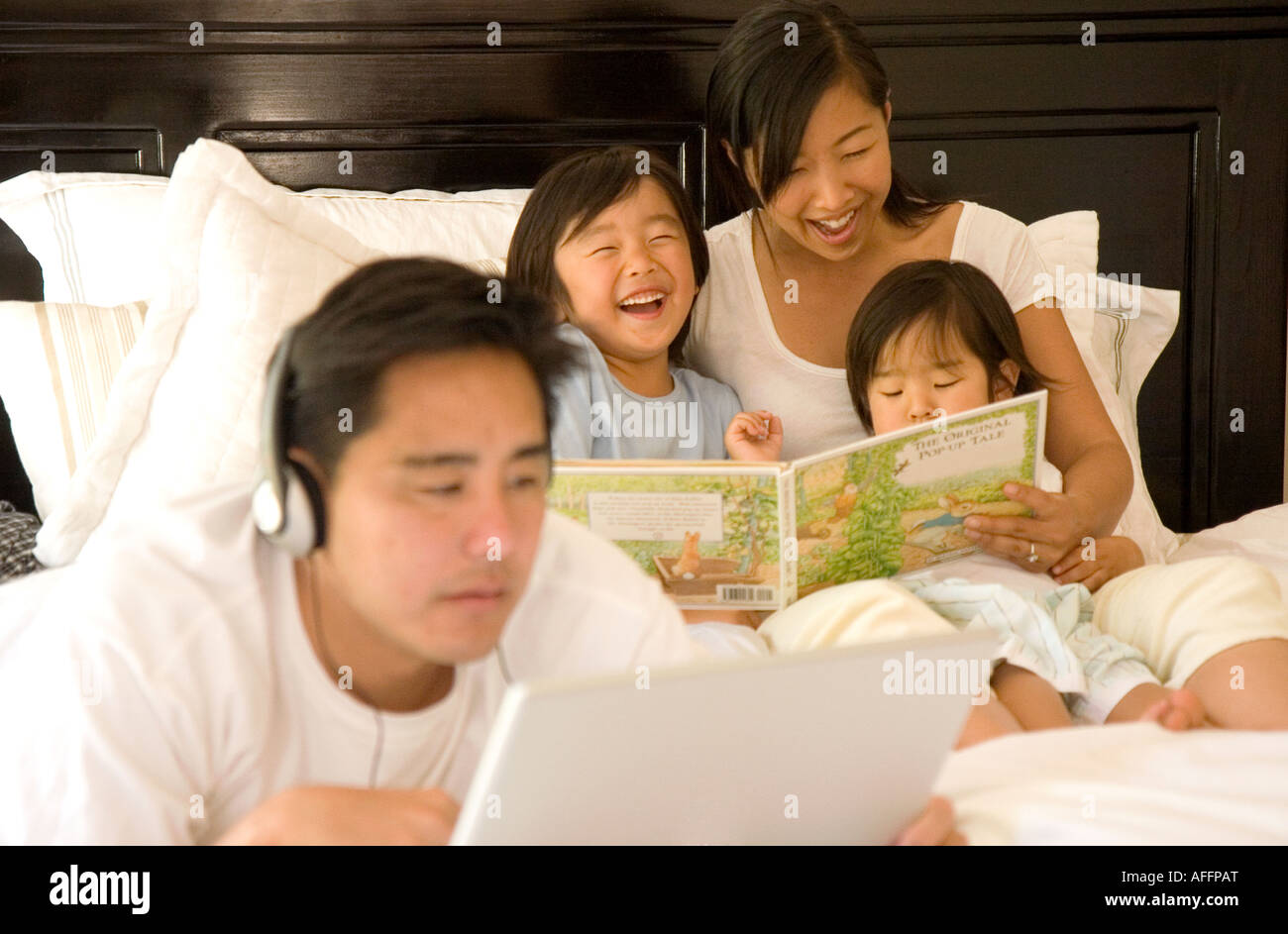 Padre ascolta le cuffie agganciata al computer mentre la madre di letture per bambini in background Foto Stock