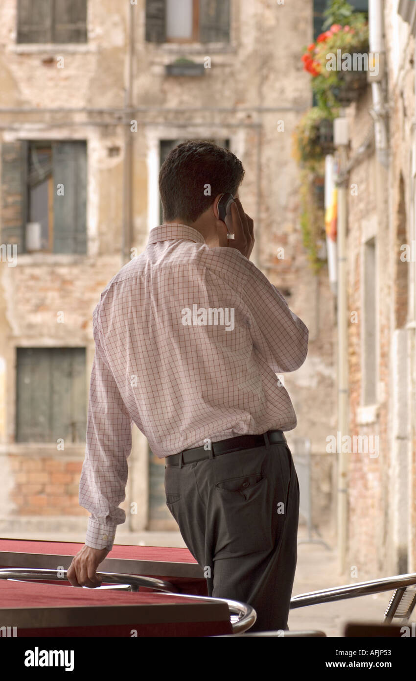 Uomo in business vestiti su un telefono cellulare visto da dietro da un cafe a Venezia Italia guardando fuori su un campo con vecchi edifici Foto Stock