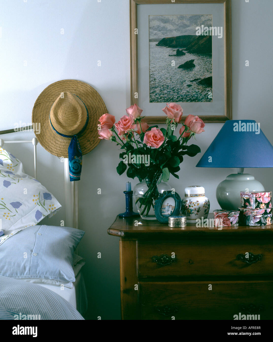 Cappello di paglia sul letto di ottone in camera da letto con seascape sopra la spia blu e rosa rose in vaso sul comodino Foto Stock