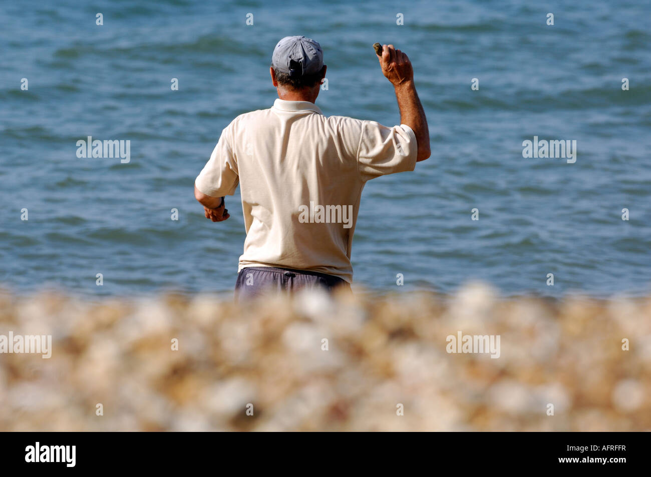 Un uomo in piedi su una spiaggia di ciottoli con il braccio alzato che lanciano pietre nel mare con l'acqua in background su una giornata d'estate Foto Stock