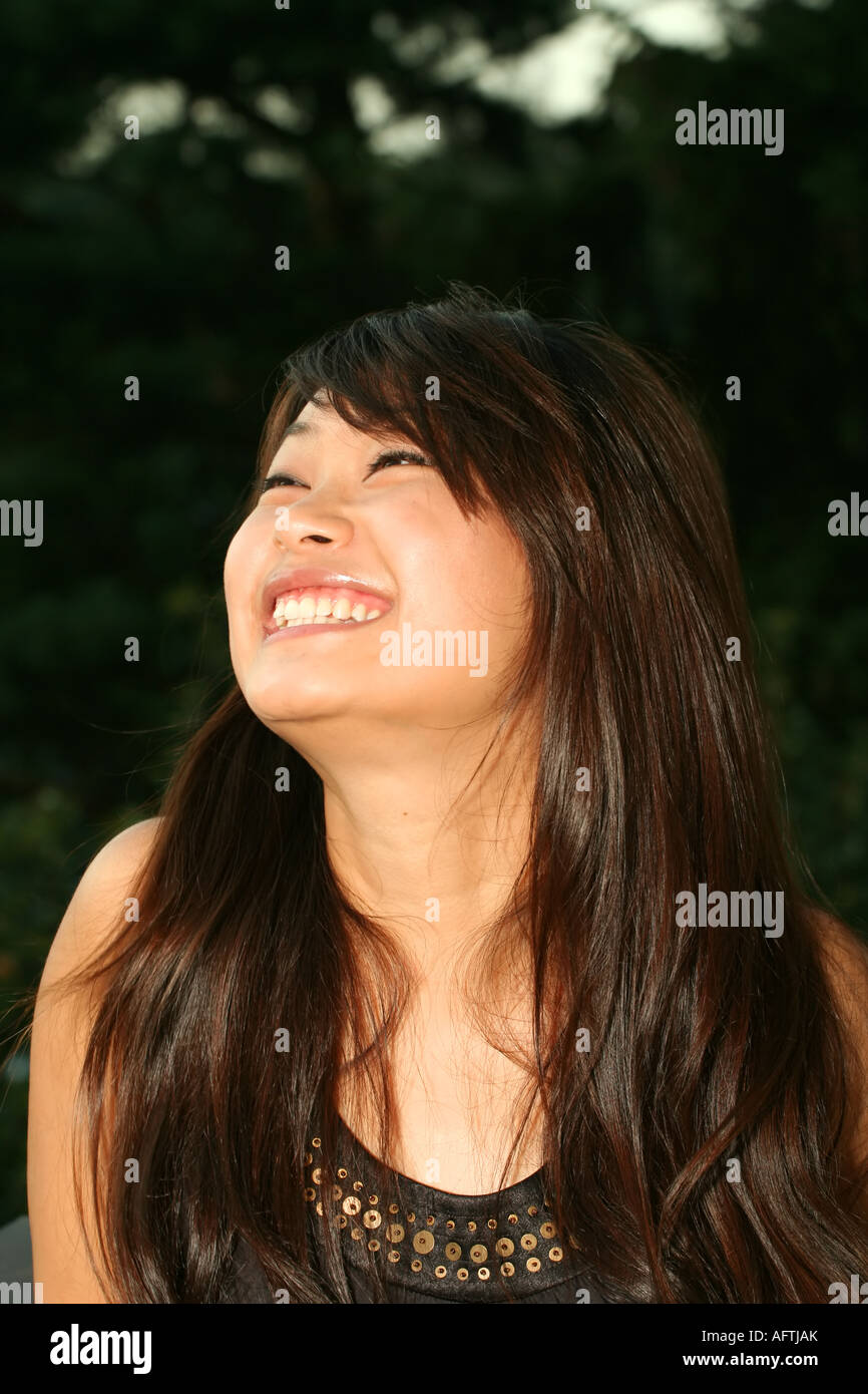 Gioiosa donna asiatica con lunghi capelli scuri, sorridente e guardando in alto, catturata all'aperto su uno sfondo verde naturale. Foto Stock