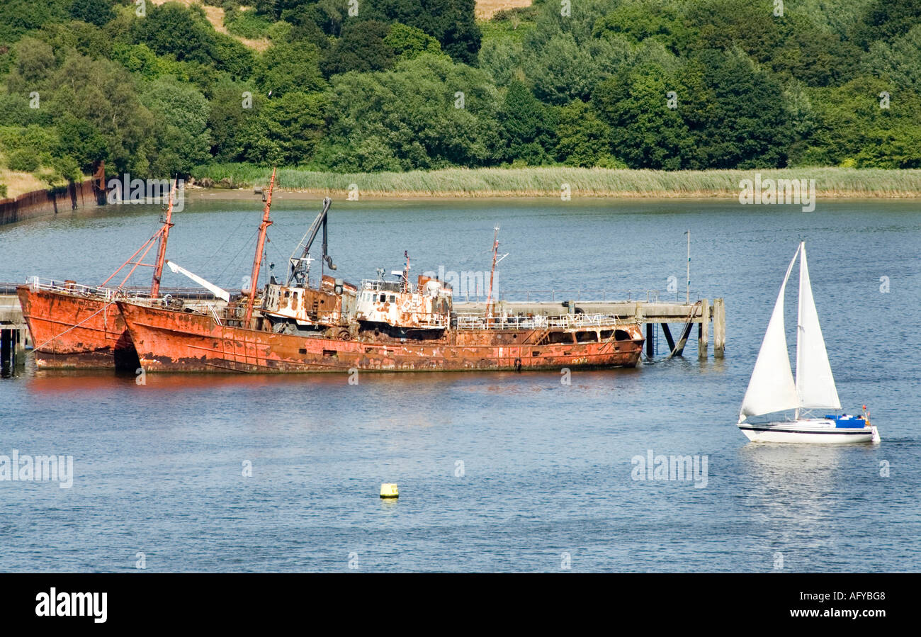 Ipswich Fiume Orwell due arrugginimento e decadenti navi accanto al molo con passaggio di barca a vela Foto Stock