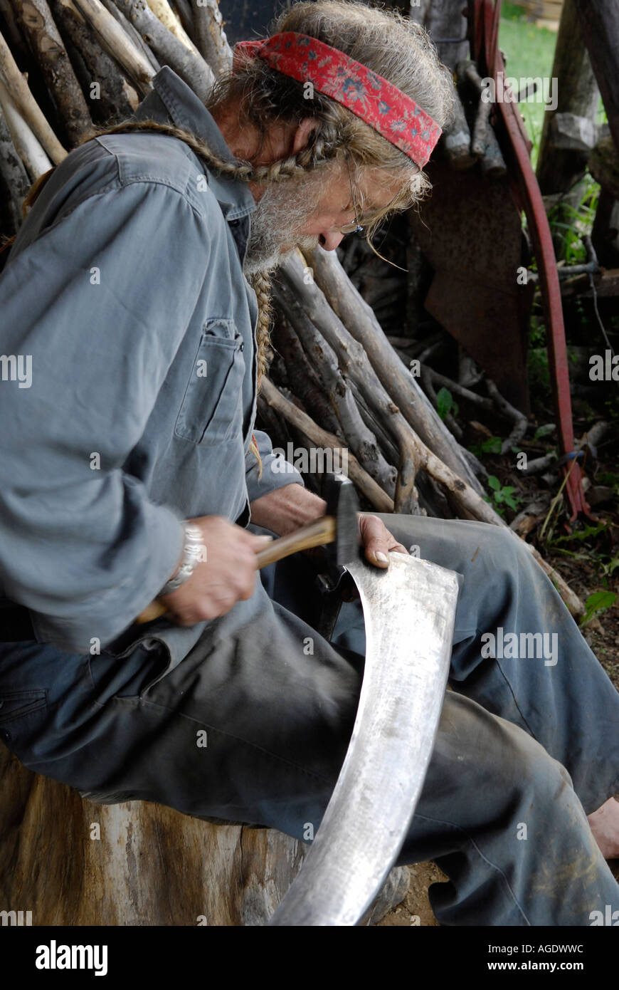 Stock a immagine di un uomo con lunghi capelli intrecciati la pallinatura e affilatura o la levigatura di una lama di falce Foto Stock