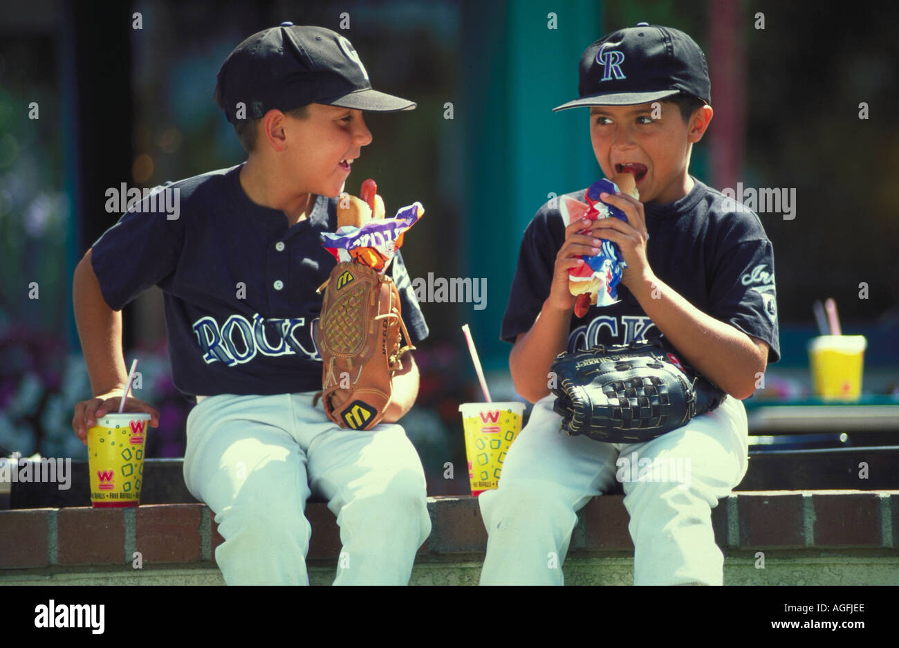 Due giovani ragazzi che indossano uniformi da baseball seduto su una parete tenendo i loro guanti mentre mangiare hot dogs e bere le acque gassate Foto Stock