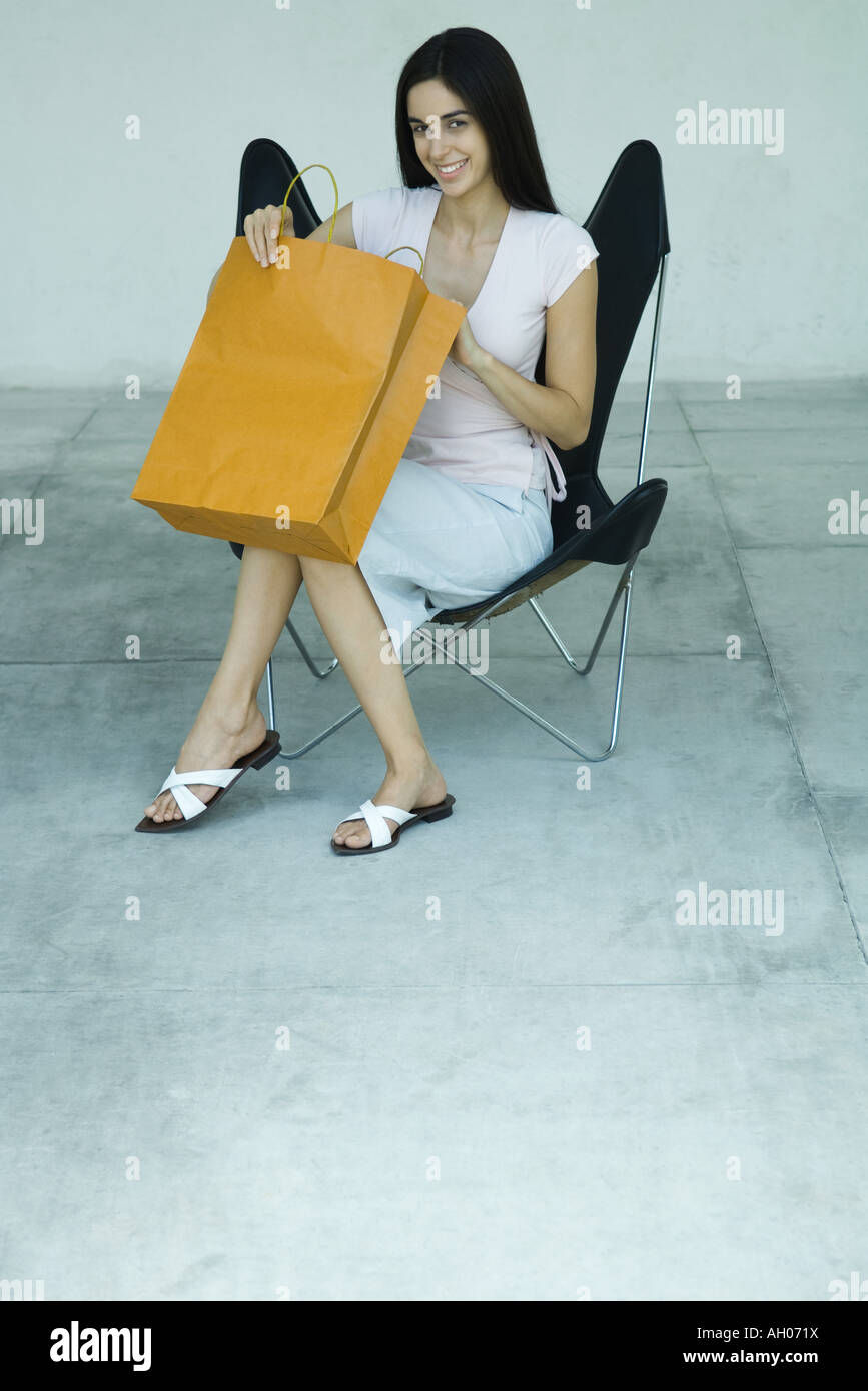 Giovane donna seduta in cattedra, apertura shopping bag, sorridente, piena lunghezza ritratto Foto Stock