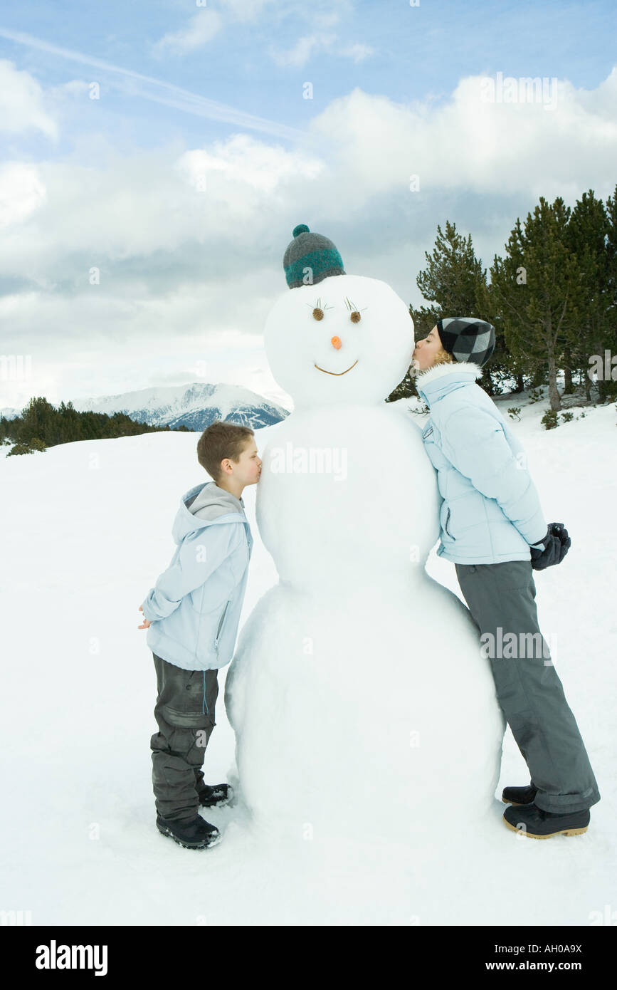 Sorella e fratello in piedi, kissing pupazzo di neve, vista laterale Foto Stock