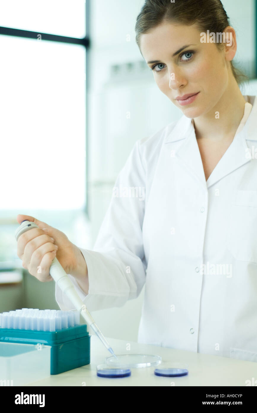 Giovane donna che lavorano nel laboratorio scientifico, tenendo un contagocce su piastra di Petri, guardando la fotocamera Foto Stock