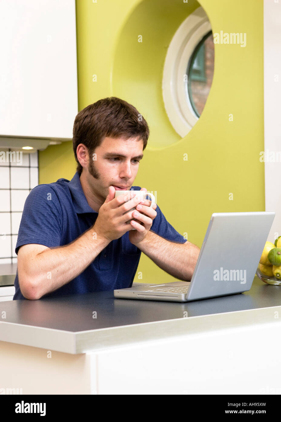 Un giovane uomo utilizza il computer in cucina mentre vi gustate una bevanda calda Foto Stock