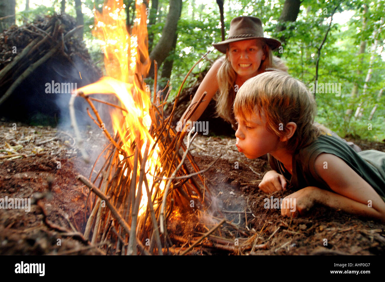 Piccolo Ragazzo in baseball hat soffia un fuoco di campo in vita in una radura del bosco come una donna guarda su approvingly Foto Stock