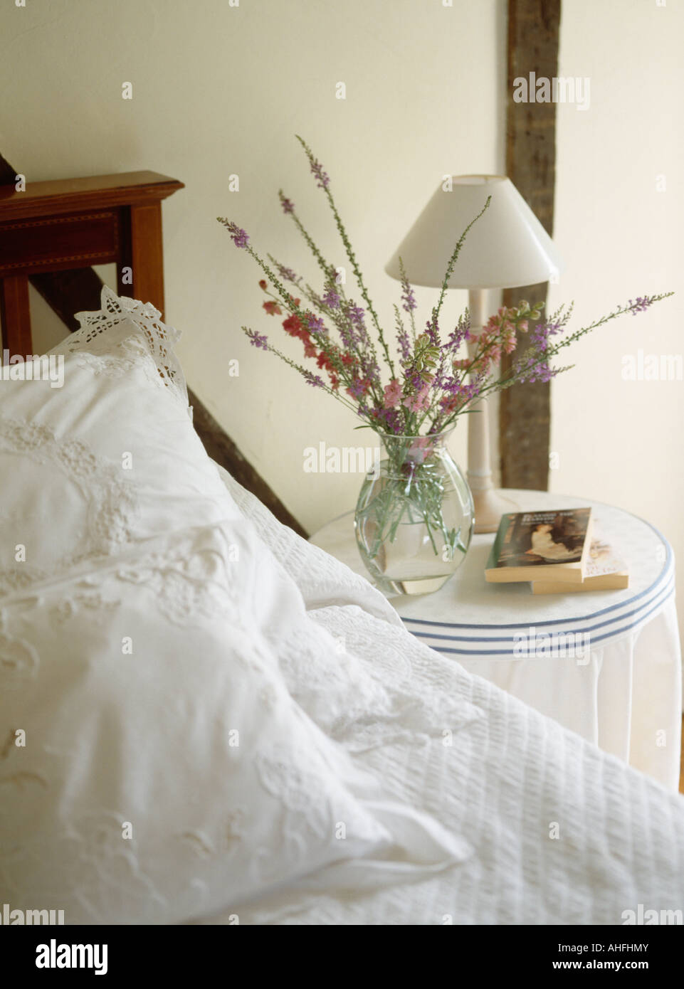 Lampada bianca e fiori freschi in vaso di vetro sul comodino accanto al letto con il bianco broderie anglaise cuscini Foto Stock