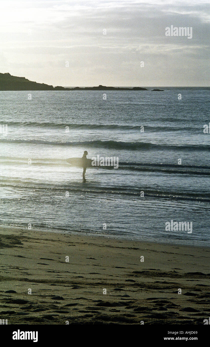 Surfista con tavola da surf in piedi, silhouette contro il sole del tardo pomeriggio riflesso dal mare a Porthmeor Beach. St. Ives, Cornovaglia, Regno Unito. Foto Stock
